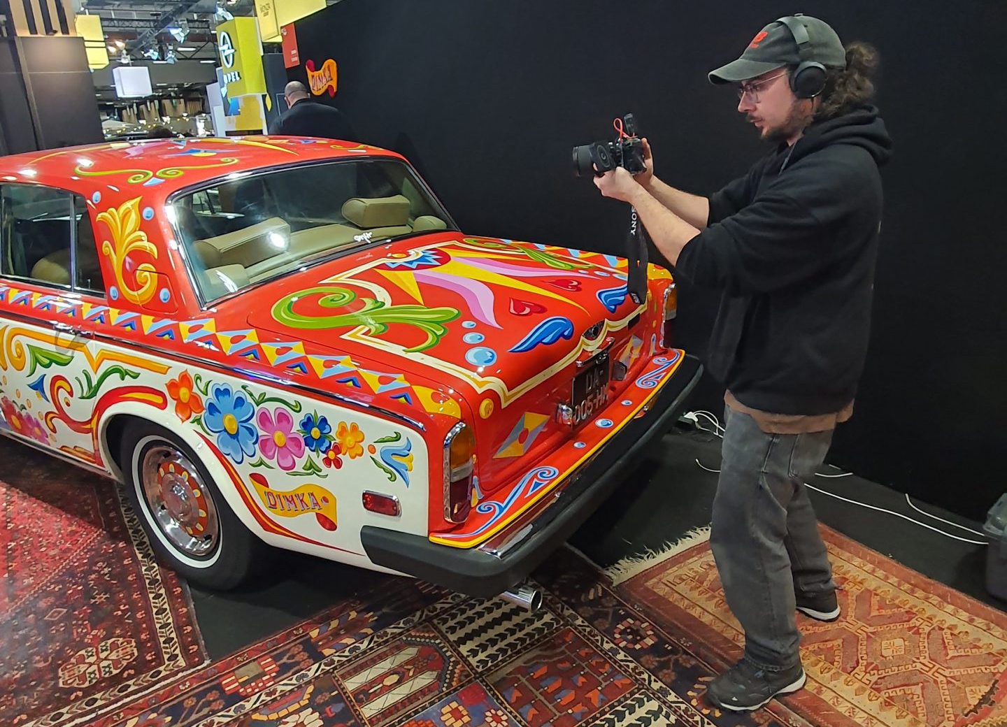 Adrien, un cadreur de l’agence vidéo Portised, filme une Bentley T2 aux couleurs psychédéliques, dans l’esprit de la Rolls-Royce de John Lennon au salon Rétromobile 2026. Made by Dimka Crafts.